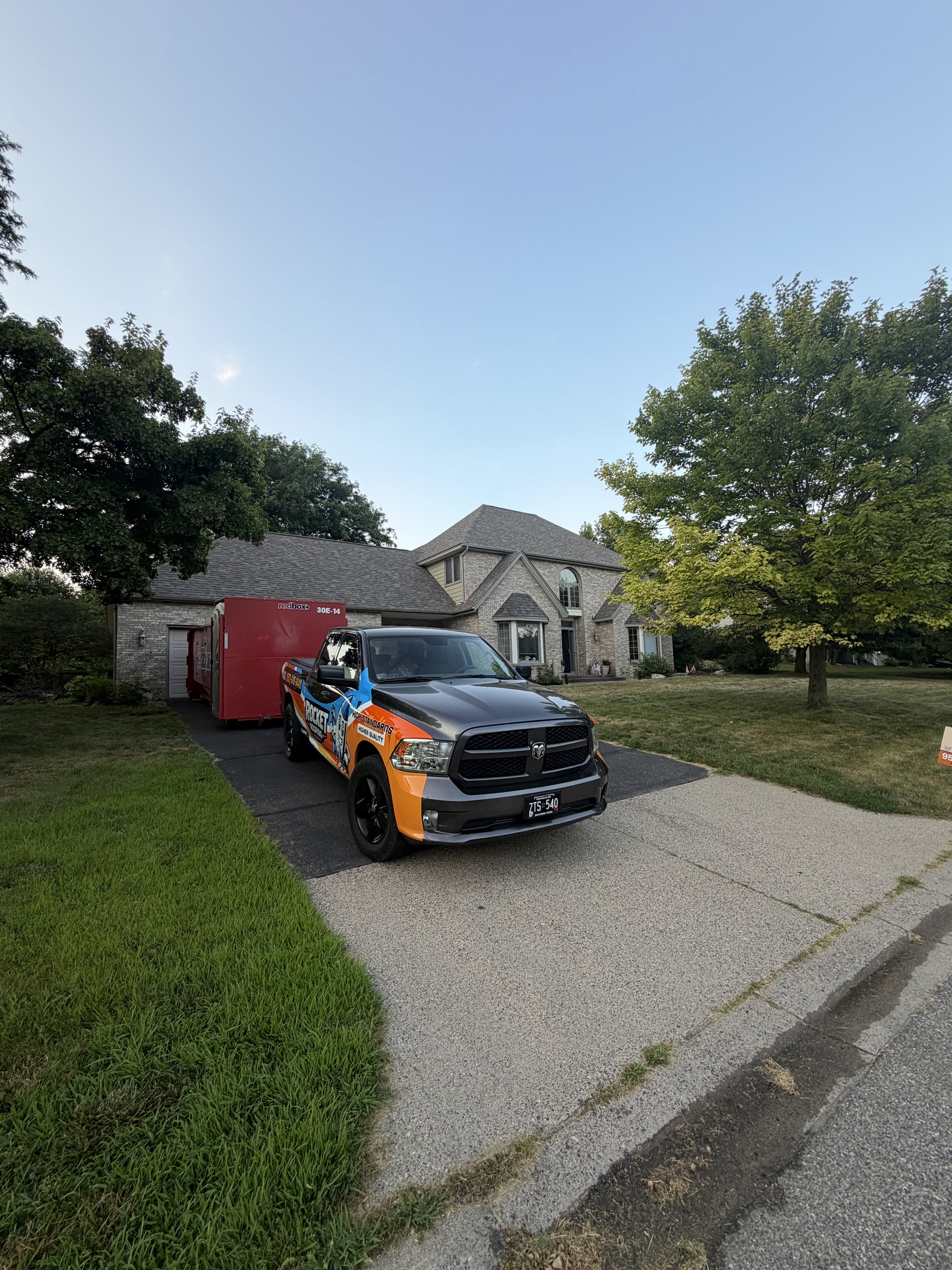 Rocket Roofing truck parked in the driveway of a west metro home at dusk during a roof replacement, dumpster staged beside the garage